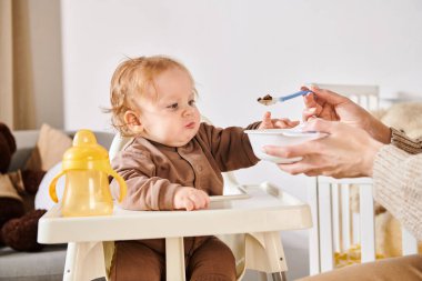 mother feeding adorable son with breakfast on baby chair in nursery room, modern parenting clipart