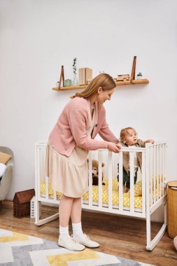 Evdeki bebek odasında beşikte duran küçük oğlunu destekleyen neşeli bir kadın. Mutlu bir annelik.