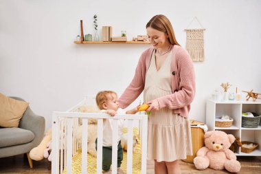 Bebek odasında beşiğinde oğluyla oynarken oyuncak tutan neşeli hamile kadın.