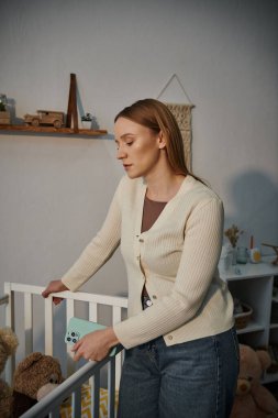 grieving young woman with smartphone standing near crib with soft toys in bleak nursery room at home clipart