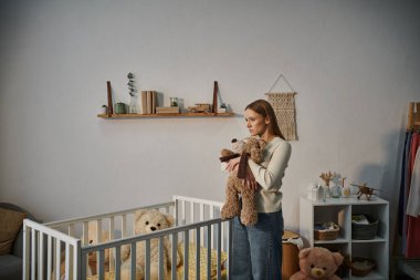 helpless and upset woman with soft toy standing near crib with in bleak nursery room at home clipart