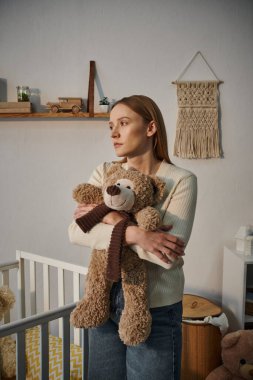 heavy-hearted grieving woman with soft toy standing near crib in bleak nursery room at home clipart
