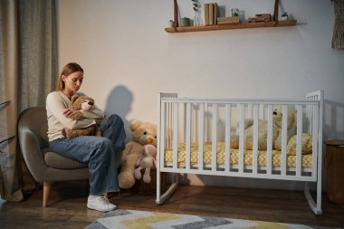 despairing young woman with soft toy sitting in armchair near crib in dark nursery room at home clipart