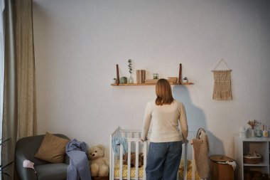 back view of depressed and lonely woman near crib with soft toys un dark nursery room at home clipart