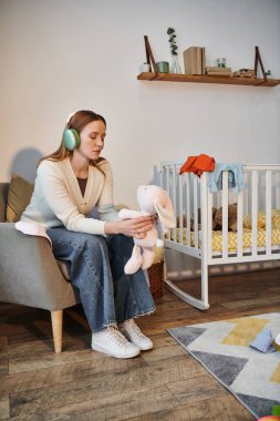 Umutsuz bir kadın elindeki yumuşak oyuncakla evdeki karanlık bebek odasında kulaklıkla müzik dinliyor.