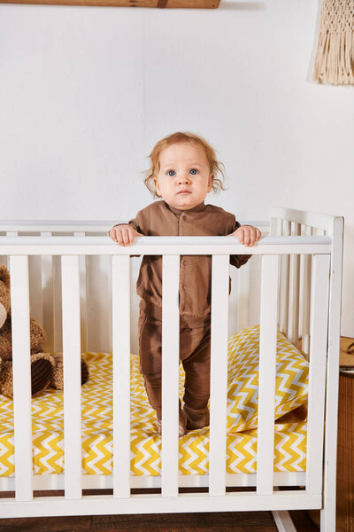 adorable toddler baby boy in romper standing in crib in nursery room at home, happy childhood