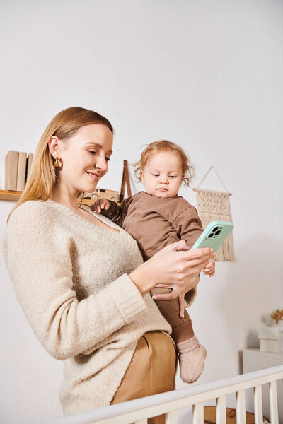 молодая мать с малышом сыном в руках сообщения на мобильный телефон в детской комнате, многозадачная женщина