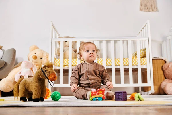 stock image little innocent boy sitting on floor near crib and soft toys in cozy nursery room, blissful babyhood