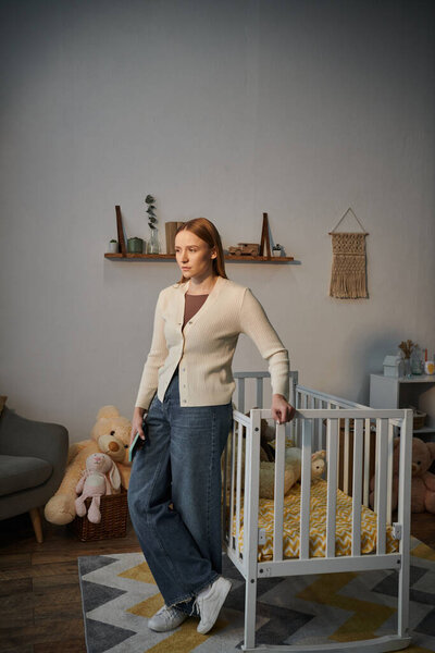 young unhappy woman standing near crib with soft toys in bleak nursery room at home, depression