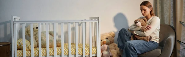 stock image depressed young woman with soft toy sitting in armchair near crib in dark nursery room, banner