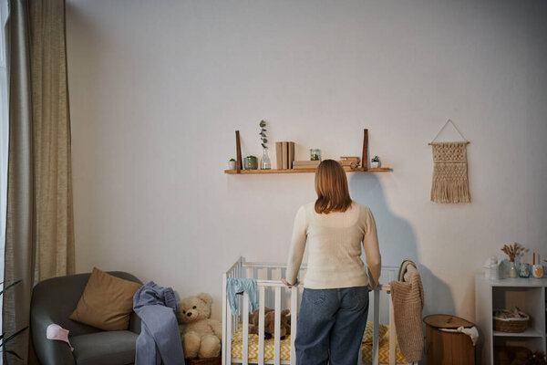 Вид сзади депрессивной и одинокой женщины возле кроватки с мягкими игрушками и темной комнаты няни дома