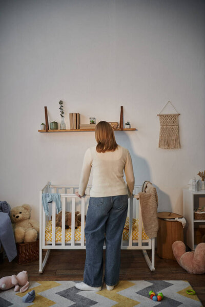 back view of grieving and depressed woman near crib with soft toys un dark nursery room at home
