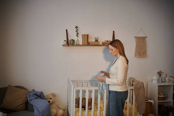 side view of frustrated woman holding baby clothes near crib with soft toys in nursery room