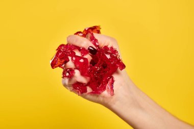 unknown young woman with nail polish squeezing red fresh jello in her hand on vivid yellow backdrop clipart