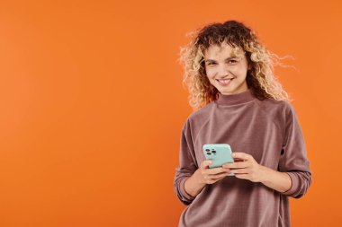 Kahverengi boğazlı kazaklı neşeli kıvırcık kadın turuncu stüdyo arka planında cep telefonuyla mesajlaşıyor.