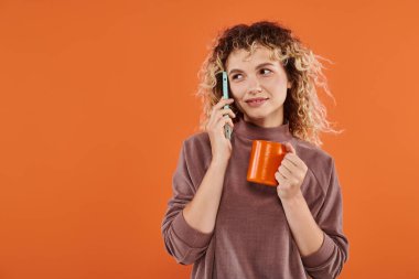 Sabah kahveli mutlu kıvırcık kadın parlak turuncu arka planda akıllı telefondan konuşuyor.