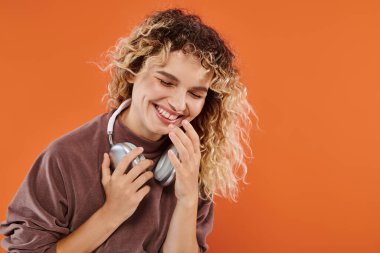 Kahverengi boğazlı, kablosuz kulaklıklı, turuncu arka planda gülen heyecanlı kıvırcık kadın.