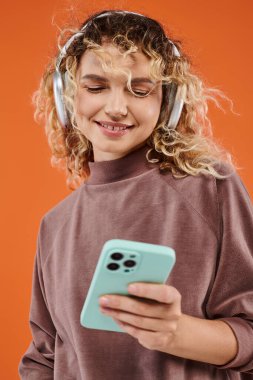 Mutlu kıvırcık kadın akıllı telefona bakıyor ve turuncu kablosuz kulaklıkla müzik dinliyor.