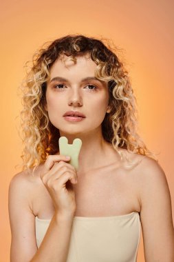 Cildi mükemmel kıvırcık, çekici bir kadın. Gua sha 'yı tutuyor. Pastel arka planda kameraya bakıyor.