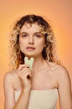 attractive curly woman with perfect skin holding gua sha looking at camera on pastel backdrop clipart