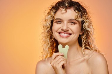 Mükemmel ciltli neşeli kıvırcık kadın Gua sha 'yı tutuyor Pastel arka planda kameraya bakıyor