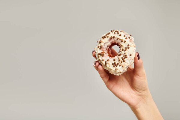 object photo of delicious donut with white icing in hand of young unknown woman on gray backdrop