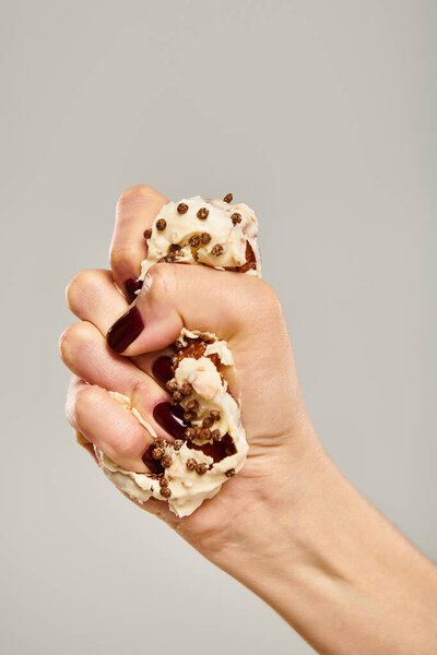 young unknown female with nail polish squeezing delicious donut with white icing on gray backdrop