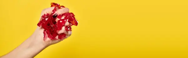 stock image unknown woman with nail polish squeezing red jello in her hand on vibrant yellow backdrop, banner