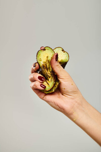 unknown female model with nail polish squeezing fresh green avocado in her hand on gray backdrop