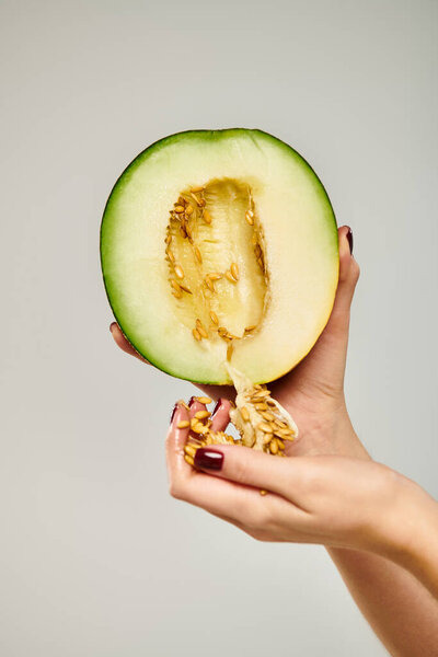 unknown woman with nail polish picking up seeds from delicious cantaloupe on gray background