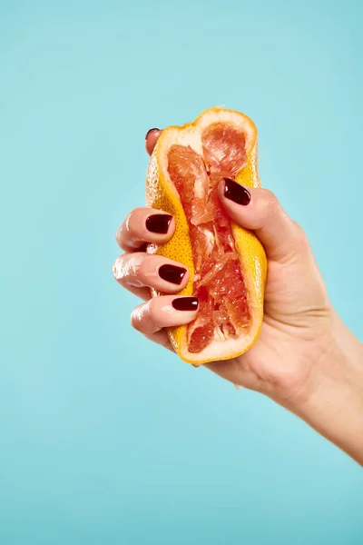 unknown woman with nail polish squeezing gourmet fresh grapefruit in her hand on blue background