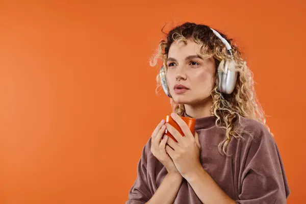 pensive curly woman with cup of morning coffee listening music in wireless headphones on orange