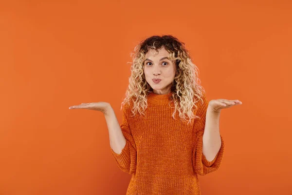 stock image confused curly woman in knitted sweater puffing cheeks and showing shrug gesture on orange