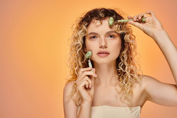 attractive curly woman with natural makeup using jade rollers looking at camera on pastel backdrop