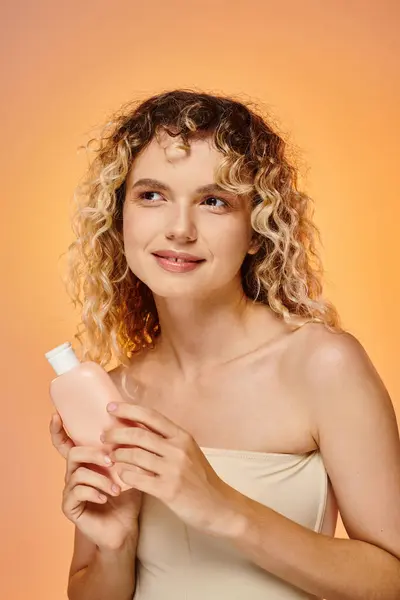 stock image cheerful woman with curly hair holding bottle with body lotion on pastel gradient backdrop