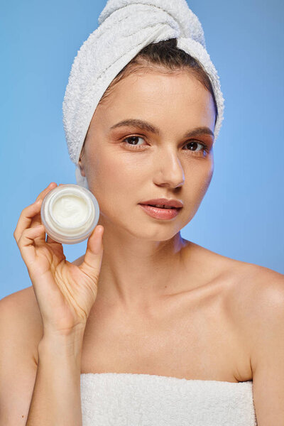 attractive woman with towel on head showing jar of face cream on blue backdrop, wellness and beauty