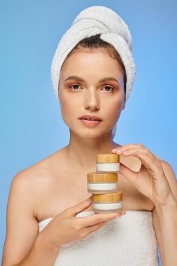 Temiz tenli ve kafasında havlu olan çekici bir kadın elinde kozmetik kremiyle mavi kameraya bakıyor.