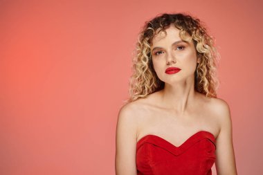 Kırmızı makyajlı şık kıvırcık kadın pembe ve sarı arka planda kameraya bakıyor.