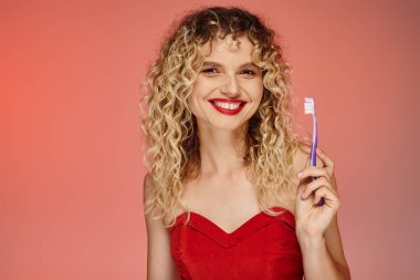 Dalgalı saçlı, kırmızı dudaklı diş fırçası tutan ve pembe kameraya bakan büyüleyici bir kadın.
