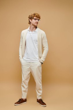 young redhead man in eyeglasses and old money style casual attire with hands in pocket on beige clipart