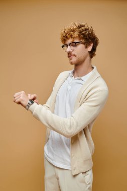 trendy redhead man in eyeglasses looking at wristwatch on beige backdrop, timeless old money fashion clipart