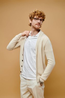 young redhead man in fashionable casual attire and eyeglasses looking away on beige backdrop clipart