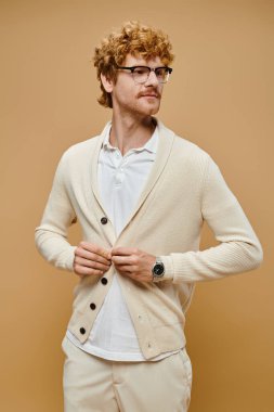young trendy male model in eyeglasses buttoning cardigan on beige backdrop, old money fashion clipart