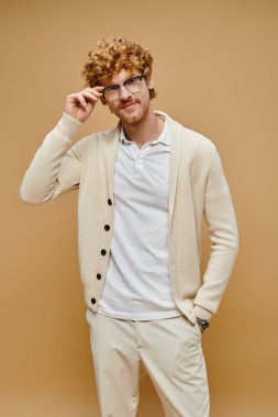 positive stylish redhead male model adjusting eyeglasses and looking at camera on beige backdrop clipart