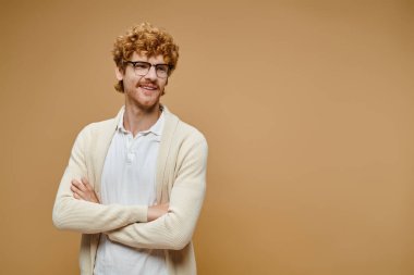 happy redhead man in eyeglasses and stylish clothes posing with folded hands looking away on beige clipart