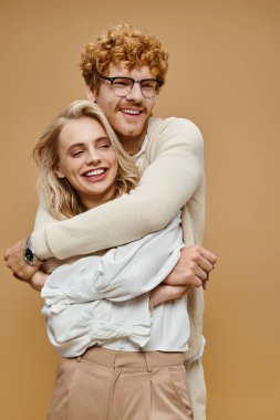 happy redhead man in eyeglasses embracing trendy blonde woman on beige, old money style couple clipart