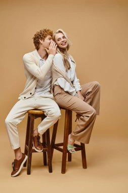 fashionable redhead man telling secret to trendy blonde woman while sitting on chairs on beige clipart