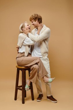 laughing blonde woman sitting on chair and hugging young redhead man on beige, old money style clipart