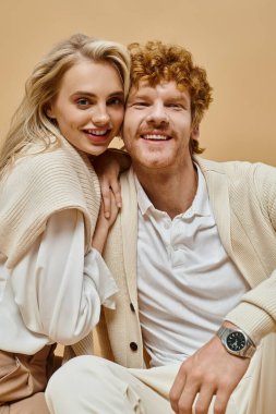 pleased blonde woman and redhead man in light-colored outfit looking at camera on beige backdrop clipart