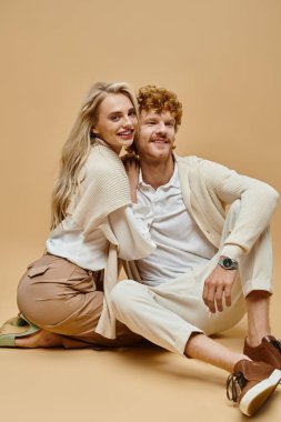 Neşeli ve genç çift, modaya uygun eski moda para kıyafetleriyle bej arka planda oturuyorlar.